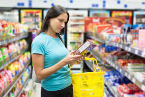 A woman with long dark hair stands in a grocery store aisle, examining a product box while holding a yellow shopping basket. Brightly colored packaging lines the shelves around her.