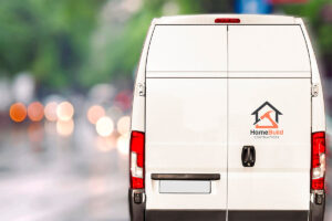 White work van with a logo reading "HomeBuild Contractors" featuring a house icon and a hammer, parked on a rainy street with blurred colorful lights in the background.