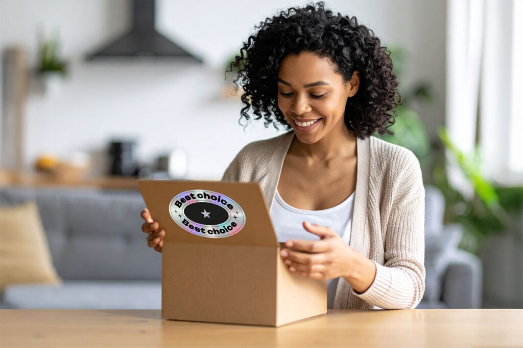 A smiling woman with curly hair opens a cardboard box on a wooden table, revealing a sticker that says "Best choice" inside. A cozy living space is visible in the background.