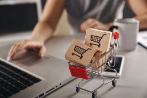 A person's hand reaching for a laptop keyboard, with a miniature shopping cart filled with small cardboard boxes featuring shopping cart icons in the foreground and a coffee mug beside it.