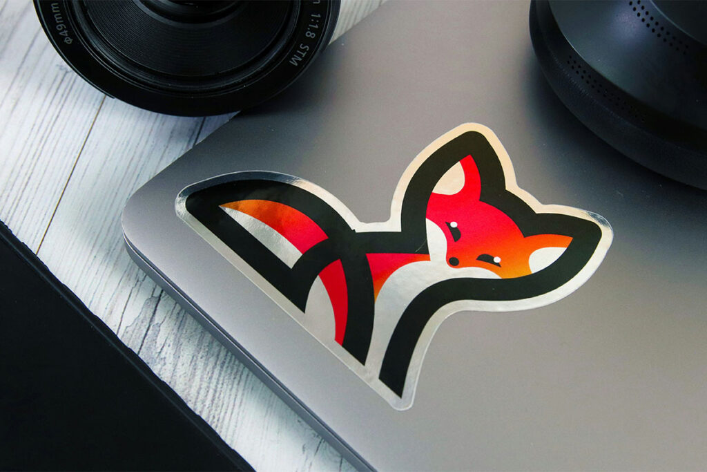 A colorful fox sticker is placed on the surface of a silver laptop, with a camera lens partially visible in the background. The laptop rests on a textured wooden surface.