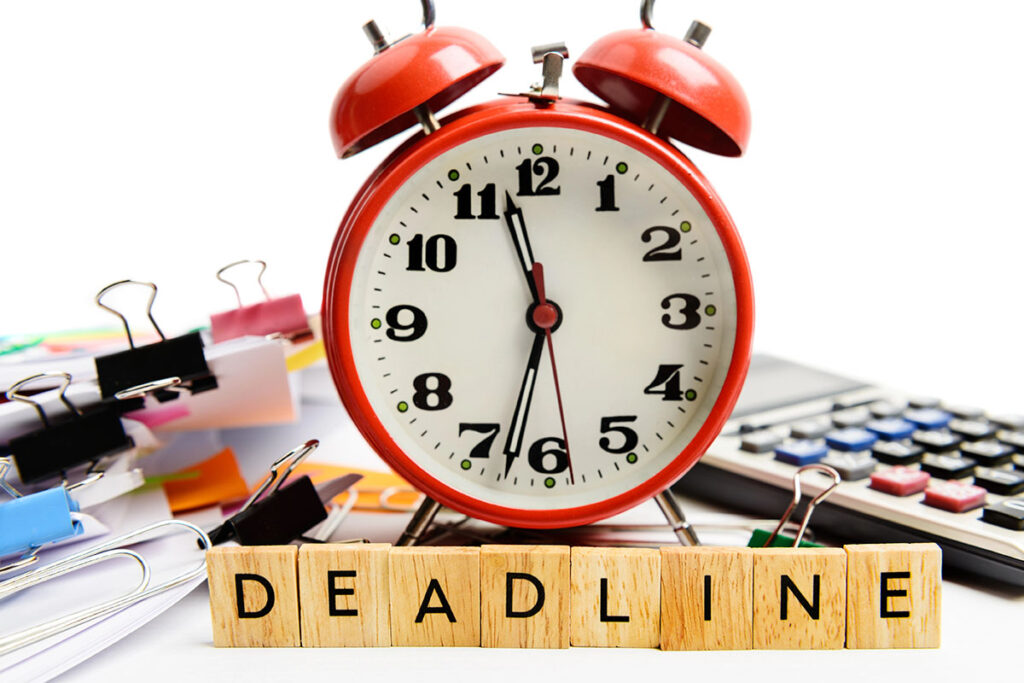 A red alarm clock showing the time around 10:10 sits next to wooden blocks spelling "DEADLINE," surrounded by paper clips and a calculator on a white background.