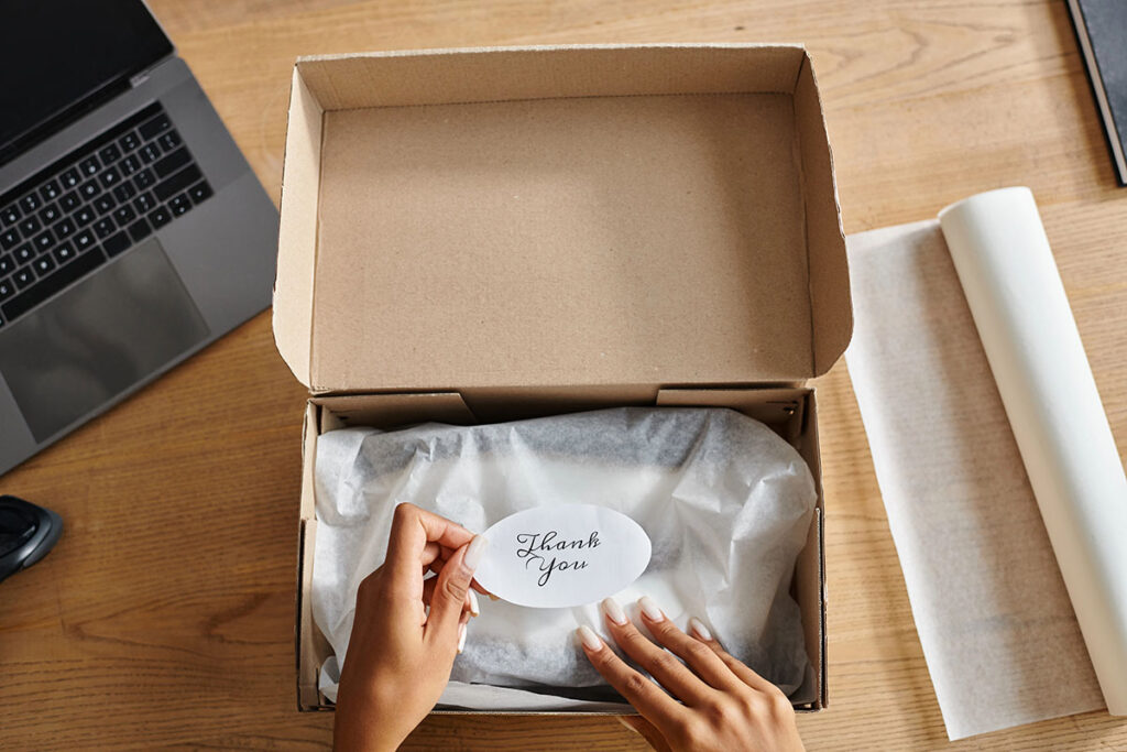 A person holds a "Thank You" card above a wrapped item inside a cardboard box, with a laptop and white paper roll visible on a wooden table.