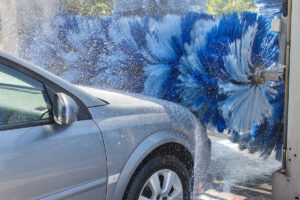 A silver car is partially washed by large blue and white brushes in an automated car wash, with water spraying around it.