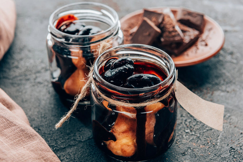 Two glass jars filled with dark syrup and whole fruits sit on a textured surface, with a piece of brown string tied around each jar. In the background, a plate holds several pieces of dark chocolate.