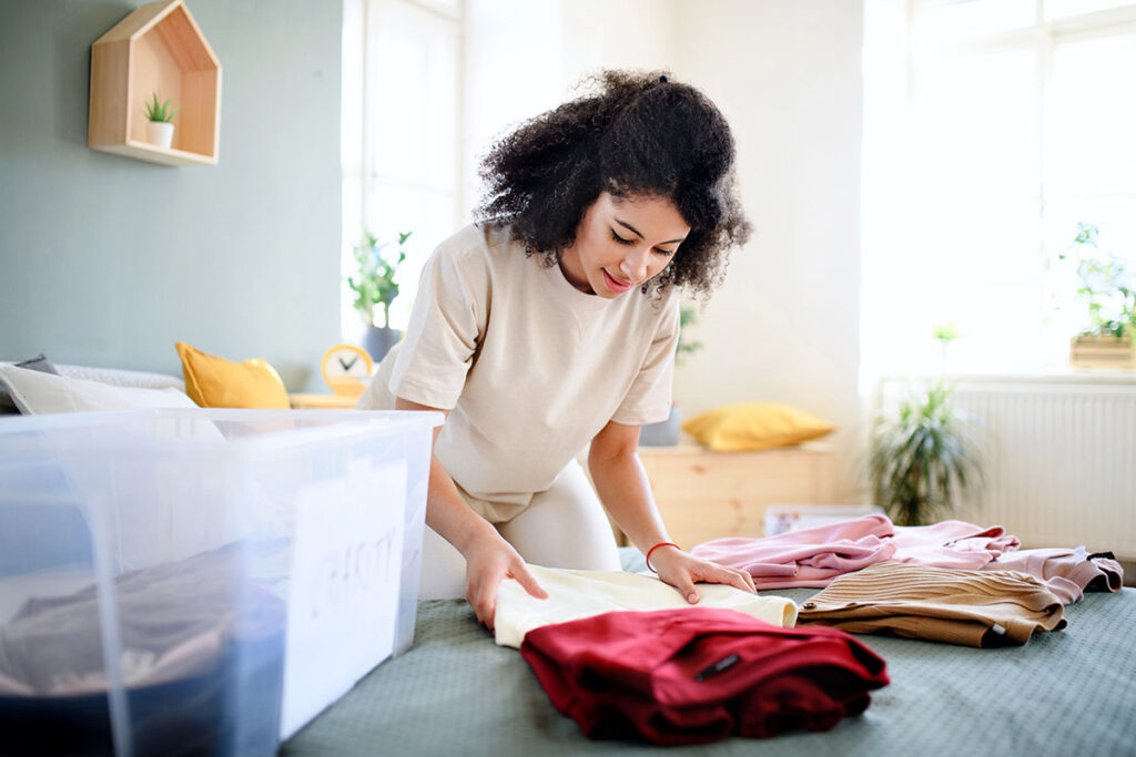 A woman with curly hair is folding clothes on a bed in a bright room, surrounded by neatly organized garments and a clear storage box. Plants and a clock are visible in the background.