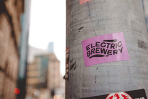 A close-up of a purple sticker on a utility pole featuring the text "Electric Brewery" in bold, black letters, with a blurred cityscape in the background.