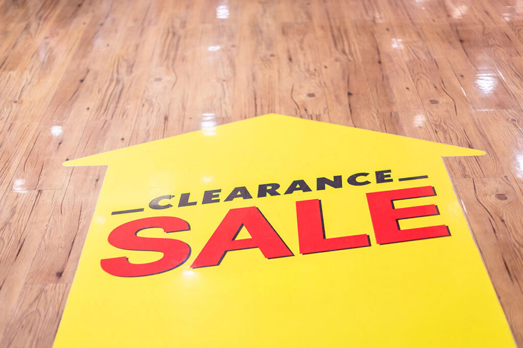 Yellow floor sticker with an arrow directing attention to a "CLEARANCE SALE" sign in bold black and red letters, on a polished wooden floor.