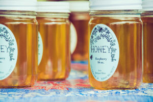 A close-up of jars of raspberry honey with decorative labels displaying the text "HONEY," "Raspberry," and "16 oz." against a colorful patterned background.