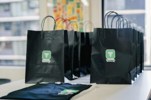 A row of black gift bags with green "Thridday" logos, neatly arranged on a table, alongside a folded black t-shirt with the same logo. Natural light filters in through a window in the background.