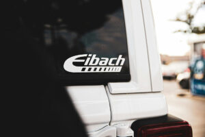 Close-up of an Eibach logo decal on the rear window of a white vehicle, with blurred cars in the background.
