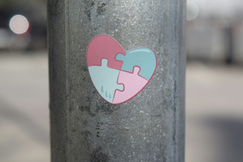 A heart-shaped sticker in shades of pink and light blue, featuring puzzle piece designs, affixed to a metal pole.
