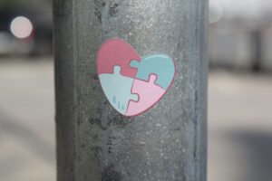 A heart-shaped sticker in shades of pink and light blue, featuring puzzle piece designs, affixed to a metal pole.