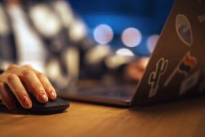 A close-up of a hand with decorated nails using a black computer mouse, with a blurred laptop covered in stickers in the background. Soft bokeh lights are visible, creating a warm ambiance.