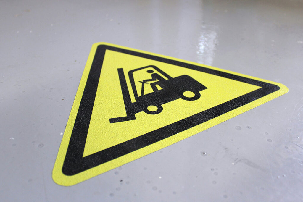A caution sign featuring a forklift and a person operating it, set against a yellow triangular background. The sign is placed on a gray floor surface.