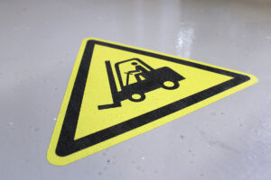 A caution sign featuring a forklift and a person operating it, set against a yellow triangular background. The sign is placed on a gray floor surface.