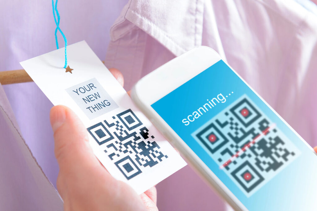 A close-up of a hand holding a clothing tag with a QR code and the text "YOUR NEW THING," next to a smartphone displaying a scanning interface.