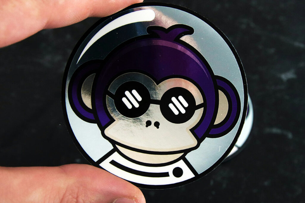 A circular metallic sticker featuring a purple cartoon monkey wearing sunglasses and an astronaut suit.
