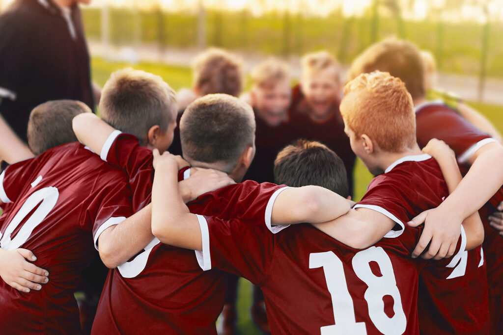 A youth sports team in matching maroon jerseys huddles tightly together outdoors with their arms around each other's shoulders.