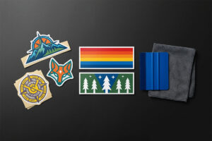 A flat lay workspace comparison showing vibrant custom die-cut vinyl decals, standard rectangular bumper stickers, a blue squeegee application tool, and a microfiber cloth laid out on a dark surface.